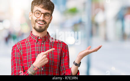 Junger stattlicher Mann mit Brille über isolierte Hintergrund erstaunt und lächelt in die Kamera während der Präsentation mit Hand und zeigte mit dem Finger. Stockfoto