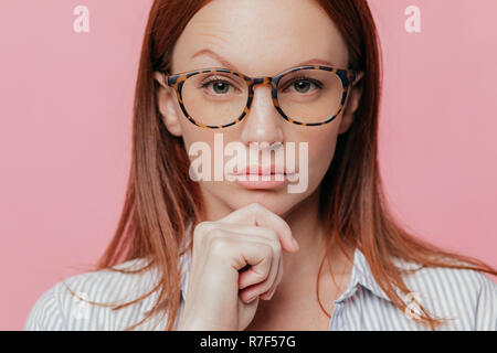 Close up Portrait von Selbstbewusste weibliche Geschäftsinhaber Kinn hält, wirft die Augenbraue, trägt optische Gläser, hat dunkle Haare, im eleganten Hemd bekleidet, Stockfoto