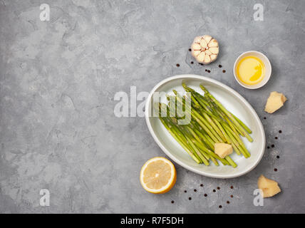 Zutaten für Backen: frischen grünen Spargel, Knoblauch, Olivenöl, Parmesan, Zitrone auf einem grauen Stein Hintergrund. Ansicht von oben Stockfoto