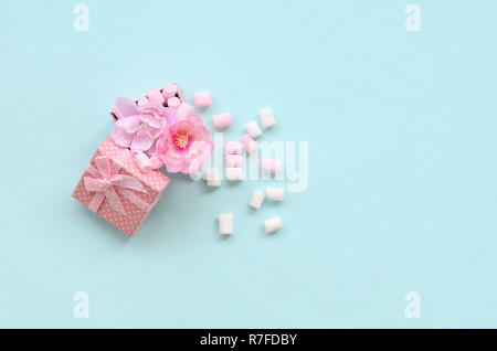 Rosa Geschenkkarton mit Blumen bestreut mit Marshmallows auf blauem Hintergrund. Metaphorische Konzept, die Verarbeitung, den Anbau von Blumen. Minimalismus flach Stockfoto