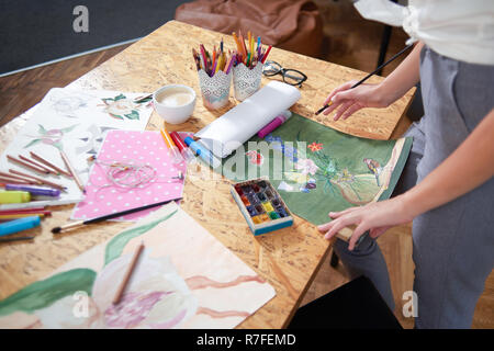 Nicht erkennbare Künstler stehen in der Tabelle mit den Maler. Maler holding Bleistift in der Hand in der Nähe von schönen Bild der Blume. Palette, Pinsel, Markierungen und Container mit Bleistift auf den Tisch. Stockfoto