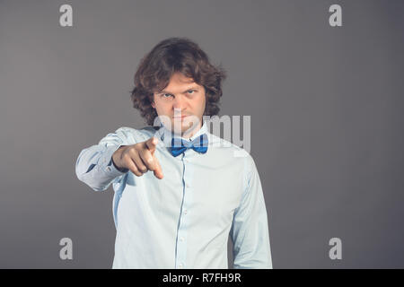 Junger Mann im blauen Hemd mit Fliege mit zotteligen Haaren, Finger direkt auf die Kamera mit dem Index Finger Pointing, tadelt sie falsche Sachen auf einem grauen Hintergrund. Hey ihr. Studio gedreht. Stockfoto
