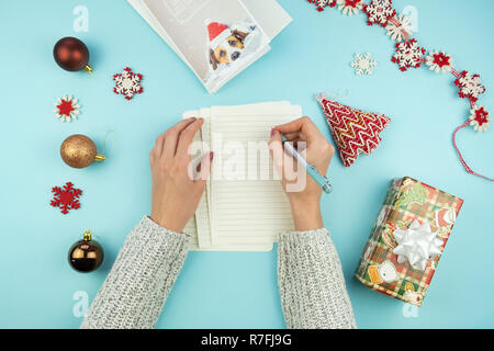 Schreiben Weihnachten Gruß schreiben. Blick von oben auf die weibliche Hände mit Stift auf blauem Hintergrund mit Papier, urlaub Requisiten und Geschenke, und das neue Jahr Karte Stockfoto