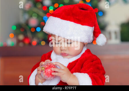 Porträt einer niedlichen kleinen Baby Boy mit Interesse erkunden Weihnachtskugel, adorable Kind Gekleidet in rot Santa Kostüm Weihnachten feiern im Stockfoto