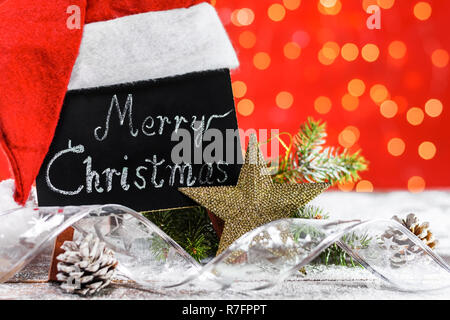 Eine Tabelle mit den Text "Frohe Weihnachten!" in einem Chalk mit Santa's Hut auf einem Holzbrett mit Schnee mit einem Zweig der Fir fallen geschrieben, Kegel Stockfoto