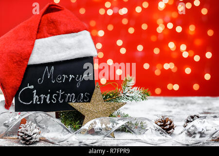Eine Tabelle mit den Text "Frohe Weihnachten!" in einem Chalk mit Santa's Hut auf einem Holzbrett mit Schnee mit einem Zweig der Fir fallen geschrieben, Kegel Stockfoto
