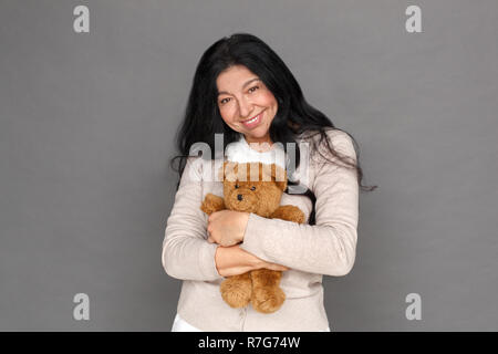 Freestyle. Reife Lady stehen isoliert auf Grau umarmen Teddybär suchen Kamera heiter Stockfoto