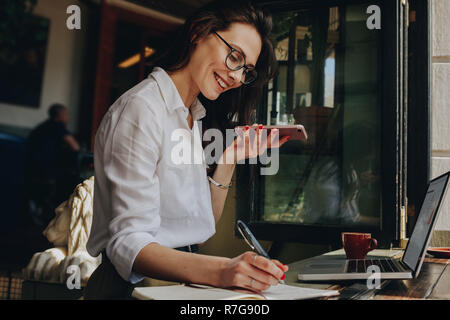 Lächelnde Frau Notizen zu machen und am Telefon sprechen während der Sitzung im Coffee Shop. Freelancer arbeiten an einem Cafe. Stockfoto
