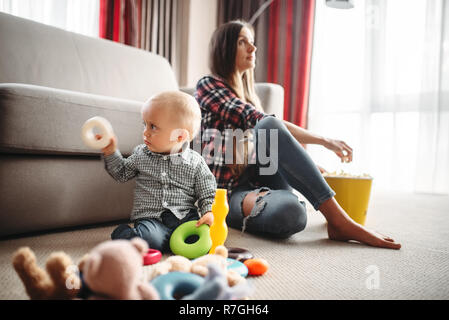 Mutter Fernsehen und isst Popcorn, wenig männliche Kind spielt in der Nähe von, Baby Care problem. Mutter und Sohn gemeinsam zu Hause glücklich, Zweisamkeit Stockfoto