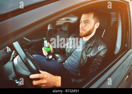 Ein betrunkener Mann, der ein Auto fährt mit einer Flasche Alkohol in der Hand. Ein Mann hält ein Antriebsrad und eine Flasche Bier. Stockfoto