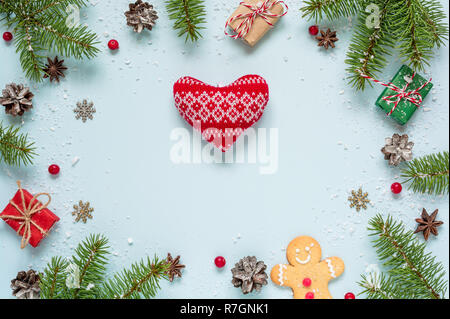 Weihnachten Rahmen aus Tannenzweigen, rote Beeren, Geschenkboxen und Tannenzapfen mit gestrickten Herz auf blauem Hintergrund. Weihnachten Hintergrund. Flach. Stockfoto