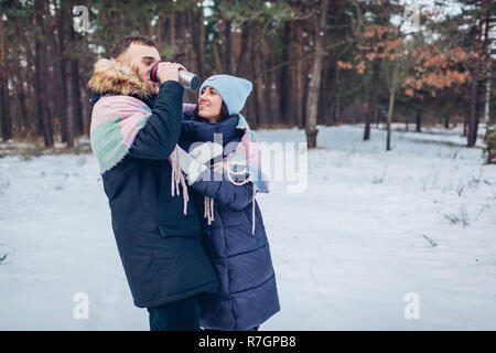 Schöne liebevolle Paar Walking im Winter Wald zusammen. Die Leute trinken heißen Tee in der Thermoskanne Schale Stockfoto