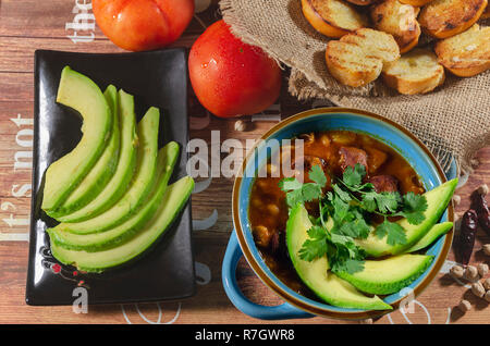 Eintopf mit Chorizo ​​and Kichererbsen, auf eine blaue Platte mit Avocado und Toast garniert mit Koriander auf einem Tisch in Low Key Licht dekoriert Stockfoto