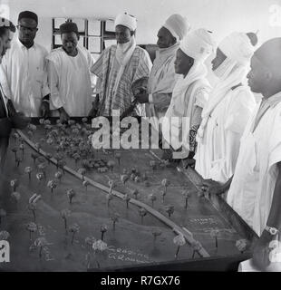 1950, historische, in einem Zimmer, einer Gruppe von nigerianischen Stammesangehörigen oder Leiter in afrikanischen Gewändern und headresses Studie ein Muster oder Modell eines Dorfes entscheiden, wie das Land verwendet werden soll ... Hier sehen wir die Schule Spielen der Felder und dem Land für die Beweidung von Vieh und Land zugewiesen wird, Nigeria. Stockfoto