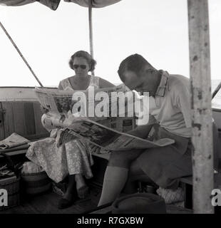 1950er Jahre, ein Mann und wpman auf einem Boot am Viktoriasee, Uganda sitzen, Zeitung lesen, der Daily Mail. Die britische Tageszeitung in dieser Ära war ein Prospekt und würde zu den britischen Kolonien und Protektoraten in Übersee, wie Uganda geschickt wurde, bevor das Land 1962 unabhängig wurde. Stockfoto