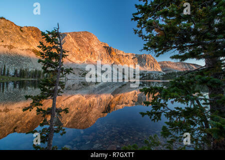 Das Licht des Sonnenaufgangs Streiks die steilen Felswände der Snowy Range in Wyoming, worin sie die noch Oberfläche eines Sees. Stockfoto