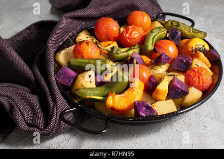 Rustikalen Ofen gebackenes Gemüse in schwarz Auflaufform Kühlung auf auf dunklen Tuch auf Hellgrau konkreten Hintergrund. Saisonale vegetarische vegane Mahlzeit. Ingredie Stockfoto