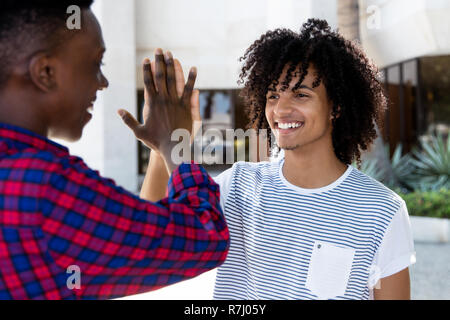 Junger Erwachsener Mann mit dem langen Haar geben hohe fünf afrikanischen Freund im Freien in der Stadt Stockfoto