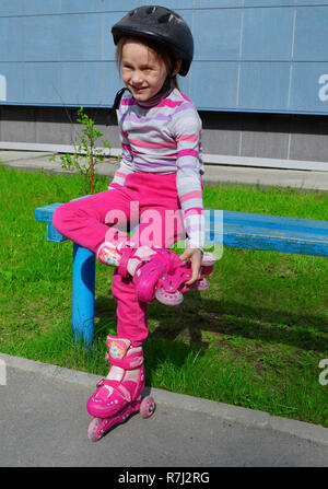 Ein Mädchen in rosa Jeans und einem gestreiften Pullover in einem Roller Helm auf dem Kopf und der Rollen auf ihren Beinen sitzt auf einer Bank an einem sonnigen Sommertag. Phot Stockfoto