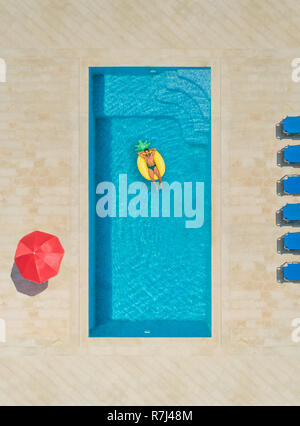 Luftaufnahme des Menschen auf Ananas geformte aufblasbare Matratze im Swimmingpool, der von Liegestühlen und Sonnenschirmen umgeben. Stockfoto