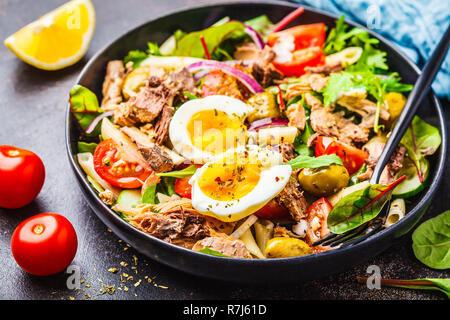 Thunfisch Salat mit Nudeln, Oliven, Gemüse und pochiertem Ei in einer schwarzen Platte auf einem dunklen Hintergrund. Stockfoto