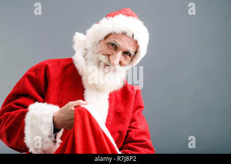 Santa Claus mit einem weißen Bart Tragen santa Outfit stehen auf dem grauen Hintergrund und zieht ein Geschenk aus dem Sack, Silvester, Weihnachten, Feiertage, Souvenirs, Geschenke, Shopping, Rabatte, Geschäfte Stockfoto