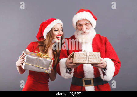 Weihnachtsmann mit weißem Bart tragen sungasses und der jungen Frau. Claus Santa Hut tragen, rotes Kleid und Sonnenbrille auf dem grauen Hintergrund, aufgeregt, Frau. Santa suchen an der Box Santa hält und lächelnd, Silvester, Weihnachten, Feiertage, Souvenirs, Geschenke, Shopping, Rabatte, Geschäfte, Snow Maiden Santa Claus, Make-up, Frisur, Karneval. Stockfoto
