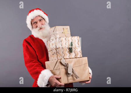 Santa Claus mit einem weißen Bart Tragen einer Brille und Santa Outfit trägt eine Menge Geschenke beim Stehen auf dem grauen Hintergrund, Silvester, Weihnachten, Feiertage, Souvenirs, Geschenke, Shopping, Rabatte, Geschäfte Stockfoto