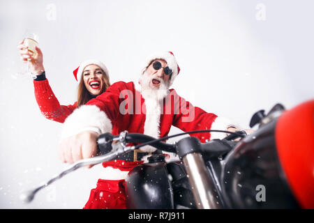 Weihnachtsmann mit weißem Bart tragen sungasses und der jungen Frau. Claus Santa Hut tragen, rot pullover und Sonnenbrille Motorrad fahren, wenn schneit, Frau Claus trinken Champagner, Silvester, Weihnachten, Feiertage, Souvenirs, Geschenke, Shopping, Rabatte, Geschäfte, Snow Maiden Santa Claus, Make-up, Frisur, Karneval. Stockfoto