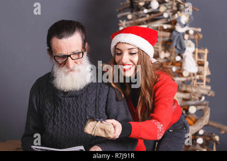 Attraktive langhaarigen Tochter im roten Pullover tragen Santa Hut gibt ein Geschenk zu Ihren bärtigen älteren Vater tragen graue Strickjacke, Silvester, Weihnachten, Feiertage, Souvenirs, Geschenke, Shopping, Rabatte, Geschäfte, Snow Maiden Santa Claus, Make-up, Frisur, Karneval. Stockfoto