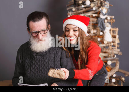 Die jungen langhaarigen Tochter im roten Pullover tragen Santa Hut lächelnd und gibt ein Weihnachtsgeschenk zu ihren bärtigen älteren Vati Brillen und graue Strickjacke, Silvester, Weihnachten, Feiertage, Souvenirs, Geschenke, Shopping, Rabatte, Geschäfte, Snow Maiden Santa Claus, Make-up, Frisur, Karneval. Stockfoto