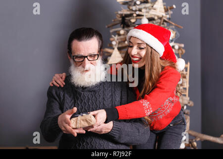 Handsom senior Vati mit weißen Bart Tragen einer Brille und graue Strickjacke akzeptiert ein Geschenk von seinem jungen Langhaarigen lächelnden Tochter im roten Pullover tragen Santa Hut, fühlte er schockiert und überrascht, Silvester, Weihnachten, Feiertage, Souvenirs, Geschenke, Shopping, Rabatte, Geschäfte, Snow Maiden Santa Claus, Make-up, Frisur, Karneval. Stockfoto