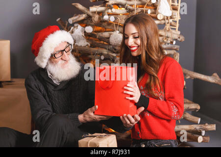 Handsom senior Vati mit weißen Bart in Brille Santa Hut und graue Strickjacke gibt ein Geschenk zu seinen jungen langhaarigen Tochter im roten Pullover, sie Uhren der Daddy Reaktion, Silvester, Weihnachten, Feiertage, Souvenirs, Geschenke, Shopping, Rabatte, Geschäfte, Snow Maiden Santa Claus, Make-up, Frisur, Karneval. Stockfoto