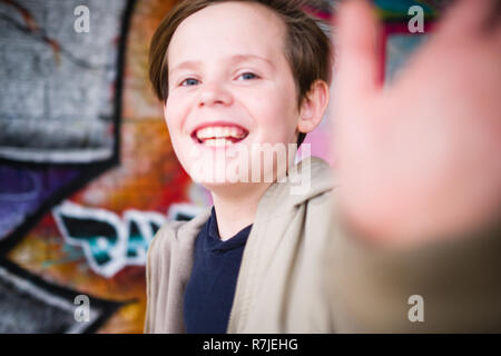 Eine lachende Kaukasischen 11 Jahre alter Junge erreicht in Richtung Kamera mit der linken Hand, während man vor einem graffitied Wand Stockfoto
