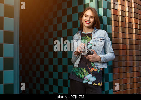 Cool Funny Girl Modell mit retro Film Kamera trägt eine Jeansjacke, dunkle Haare im Freien über Stadtmauer in einem Käfig Hintergrund. Sun flare Stockfoto