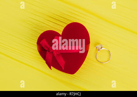 Roten herzförmigen Geschenkbox und einen goldenen Ring. Verlobungsring und Samt, in die Form eines Herzens auf gelben Hintergrund. Romantisches Geschenk Konzept. Stockfoto