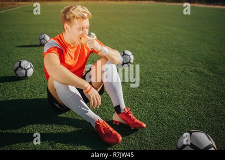 Hungrigen jungen Spieler auf dem Rasen sitzen und essen von Apple. Er hat Ruhe. Drei Kugeln sind hinter ihm. Vierte ist vor. Sonniges Wetter draußen. Stockfoto