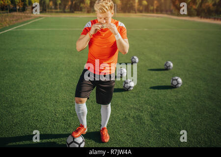 Hungrigen jungen Mann essen Sandwich. Er stand auf der grünen Wiese und halten Sie die Füße auf dem Ball. Fünf Kugeln sind hinter ihm. Sonnige Wetter draußen. Stockfoto