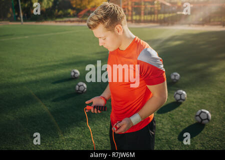 Junger Mann im Sport einheitliche bereit ist mit Seil springen zu üben. Er schaut nach unten. Kerl stehen auf der grünen Wiese. Fünf Fußball-Bälle sind hinter sich. Stockfoto