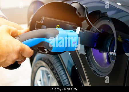 Man einschalten Laden von Auto. EV Auto oder Elektroauto an der Ladestation mit Netzkabel Netzteil in unscharfer Natur mit sanften Licht ba angeschlossen Stockfoto