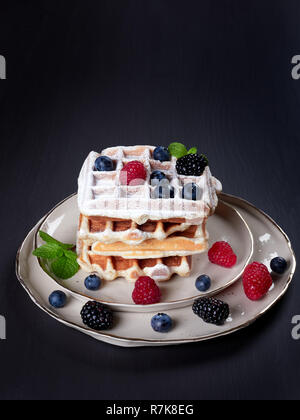 Ein Stapel mit vier home Belgische Waffeln mit Puderzucker bestäubt, mit frischen Beeren und Minze, auf eine beige Schüssel, auf dunklen braunen Hintergrund vorgenommen Stockfoto