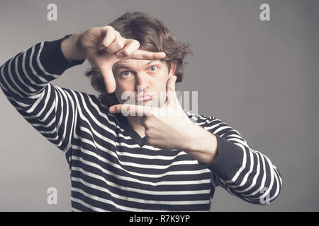 Junge schöne shaggy curly Mann, die sich mit den Fingern, Rahmung mit Händen auf grauem Hintergrund, kopieren. Nahaufnahme, Porträt Mann im gestreiften Pullover seine Hände hielten in einem framing Geste. Stockfoto