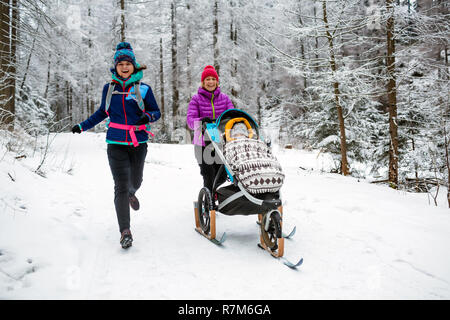 Mutter mit Baby Stroller genießen Winter Wald mit Freundin oder Partner, Zeit mit der Familie. Wandern und Laufen Frau mit Schlitten pram, Teamarbeit conce Stockfoto