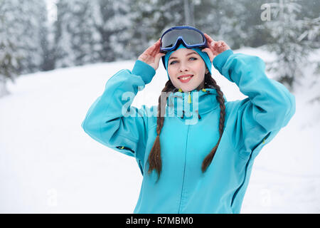 Lächelnd nordic Frau mit Zöpfen setzt an schützenden Skibrillen. Snowboarder Mädchen berühren Maske bei Ski Resort auf Schneefall in der Nähe von Forest. Blue eyed elegante Sportlerin in bunten modische Outfit. Stockfoto