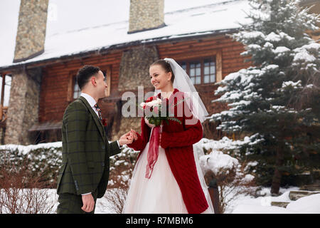 Stilvolle Paar am Hochzeitstag niedlich. Braut und Bräutigam zum ersten Mal treffen. Ersten Blick. Winter Hochzeit auf Schneefall mit schönen Holz- Ferienhaus Chalet auf Ski Resort. Frau trägt Strickjacke Stockfoto