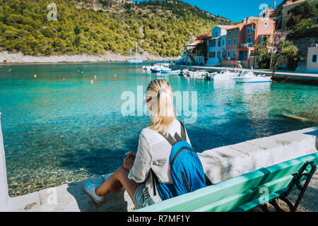 Frau Touristen sitzen auf der Bank vor der wunderschönen Bucht auf Sommermorgen. Schöne Ferienhäuser in Assos Dorf mit schönen traditionellen Häusern und pine Nut. Kefalonia, Griechenland Stockfoto