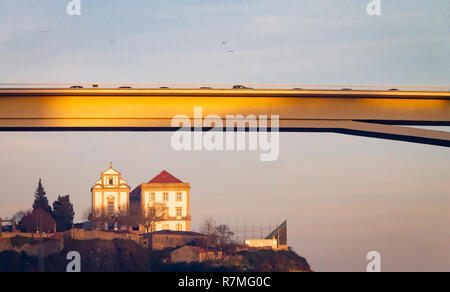 Eine Kirche ist in einem Abstand gesehen, unterhalb der Brücke Ponte Infante, in Porto, Portugal, während der Goldenen Stunde, während die Möwen von Fliegen. Stockfoto