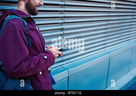 Zuversichtlich bärtigen Mann hält im Handy und während in der Nähe der Straße Wand im sonnigen Sommer Abend stehen. Stockfoto