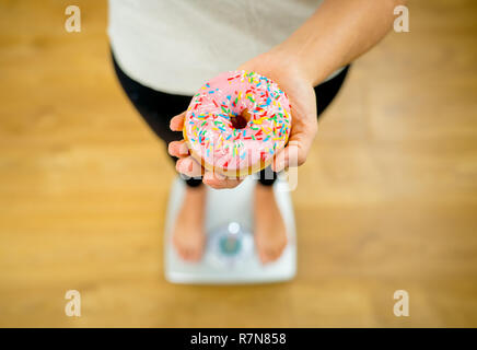 Nahaufnahme von Frau auf Skala holding Donut an Hand unglücklich von Körpergewicht und Essgewohnheiten zu viel Junk ungesunde Lebensmittel auf Diät in der Ernährung Deto Stockfoto