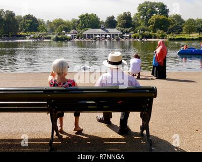 Senior Paar auf der Bank durch die Serpentine, Hyde Park, London sitzen im Sommer mit Frau im hijab im Hintergrund stehend an Water's Edge Stockfoto